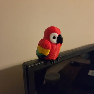 May include: A small, three-dimensional parrot figurine featuring a bright red head and body, white eye patches, a black beak, and wings colored with yellow and green accents. The parrot is perched on the top edge of a black monitor frame.