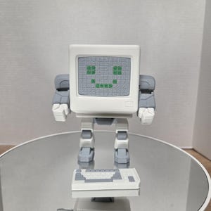 May include: A white and gray robot figurine shaped like a vintage computer monitor with a green pixelated smiley face on the screen. The robot has articulated arms and legs and is accompanied by a small, separate white and gray computer keyboard.