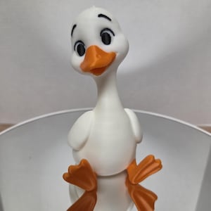 May include: A white duck figurine with a smooth finish, featuring a bright orange beak and matching orange webbed feet. The duck has large, expressive black eyes with defined eyebrows, a long neck, and a rounded body, standing in an upright position.