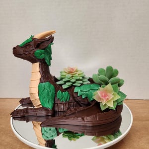 May include: A brown dragon-shaped planter with green leaf accents and tan underbelly. The dragon holds several small succulents in its back. The planter sits on a white, round, mirrored surface.
