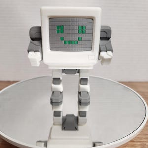Pode incluir: Uma figura de robô branca e cinza com um rosto de tela de computador. A tela tem um design pixelado com olhos verdes e um sorriso. O robô tem braços e pernas articulados e está em uma superfície reflexiva.