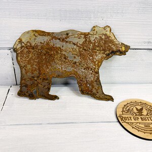 Puede incluir: Silueta de oso de metal oxidado con acabado desgastado. El oso está de pie sobre sus cuatro patas. Una posavasos de madera con el texto "Rust Up Buttercup" está al lado del oso.