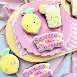 Op de afbeelding: Een verzameling versierde koekjes in de vorm van citroenen en limonade. De koekjes zijn geel, wit en roze, met de tekst "Squeeze me, I'm yours" in roze glazuur. Gerangschikt op roze en gele borden.