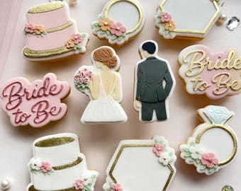 May include: A collection of decorated sugar cookies with a wedding theme. Cookies include a two-tiered pink cake, diamond rings, a bride and groom, and cookies that say "Bride to Be". The cookies are decorated with gold and floral accents.