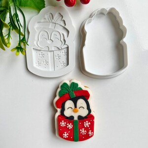 May include: A Christmas-themed cookie cutter set featuring a penguin in a gift box design. The set includes a white silicone mold and a matching cutter. A decorated cookie shows a penguin in a red gift box with white snowflakes and a green bow.