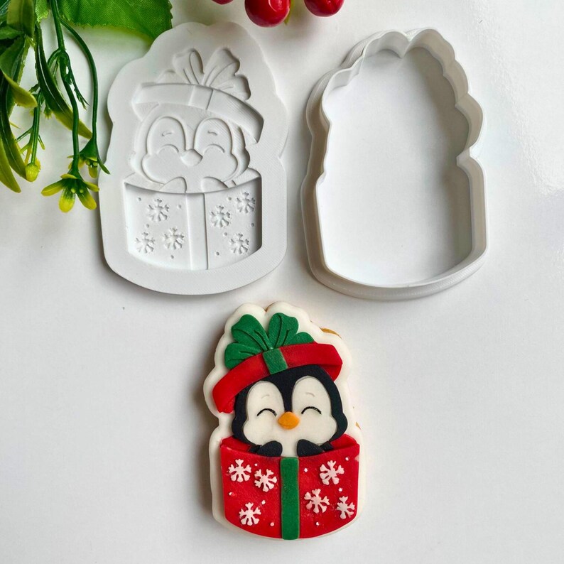 May include: A Christmas-themed cookie cutter set featuring a penguin in a gift box design. The set includes a white silicone mold and a matching cutter. A decorated cookie shows a penguin in a red gift box with white snowflakes and a green bow.