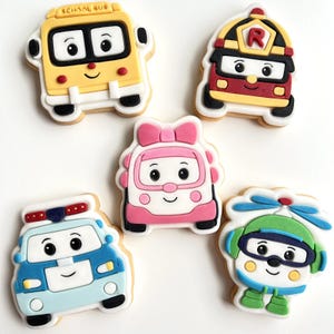 Peut inclure: Un ensemble de cinq biscuits en forme de véhicules. Ils comprennent un bus scolaire jaune, un camion de pompiers rouge, une voiture rose avec un nœud, une voiture de police bleue et un hélicoptère vert. Chaque biscuit a un visage souriant.