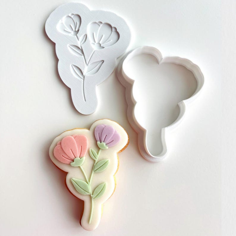 May include: A white plastic flower-shaped cookie cutter and a matching stamp are displayed next to a decorated cookie. The cookie features pink and purple flowers with green leaves on a white background. The cookie cutter is approximately 4 inches in height.