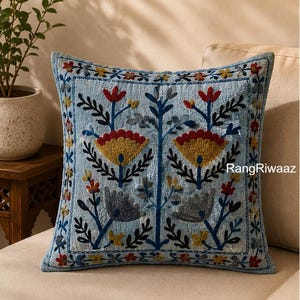 May include: A square light blue throw pillow featuring intricate multi-colored floral embroidery in red, yellow, and grey. The design includes symmetrical botanical patterns and a decorative border. The text RangRiwaaz is printed on the right side.