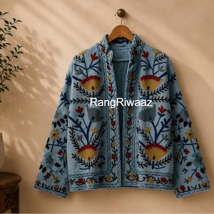 Puede incluir: Una chaqueta azul claro abierta por delante con intrincados bordados florales en amarillo, rojo y azul oscuro. La prenda muestra el texto RangRiwaaz en la parte delantera, tiene mangas largas, cuello alzado y una silueta recta estructurada.