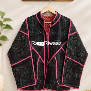 May include: A black quilted jacket with pink trim and white stitched floral patterns. The jacket features a mandarin collar and long sleeves. The interior lining is red with white polka dots. A white tag displays the text Ra Riwaaz JK 525.