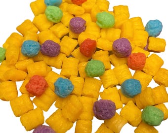 May include: A close-up of a pile of colorful cereal. The cereal is mostly yellow, with some red, blue, green, and purple pieces. The cereal is in the shape of small squares and balls.