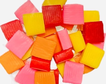 May include: A pile of colorful square candies in shades of pink, yellow, orange, and red. The candies are arranged in a random pattern and are all the same size.