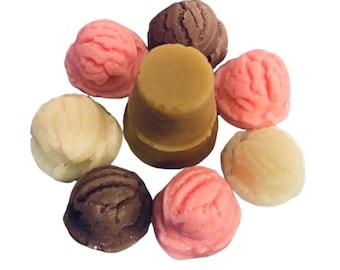 May include: An arrangement of colorful, round, ice cream-like soaps. The soaps are in shades of pink, brown, and cream, surrounding a cylindrical, light brown soap. The soaps are arranged on a white background.