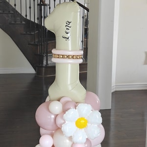 Può includere: Un grande palloncino a forma di numero uno color crema con il nome "Lena" in corsivo nero. Il palloncino è decorato con accenti dorati e rosa e poggia su un gruppo di palloncini rosa, bianchi e color crema e un grande fiore bianco con un centro giallo.