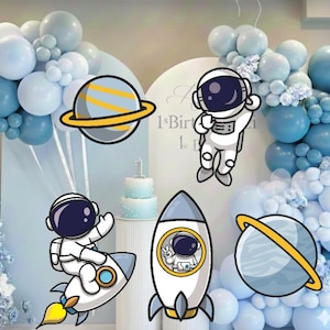 May include: A festive birthday scene with blue and white balloons, cartoon astronauts, planets, and rockets. A two-tiered cake is on a pedestal. The text "1st Birthday" is visible on the backdrop. The overall theme is space and birthdays.