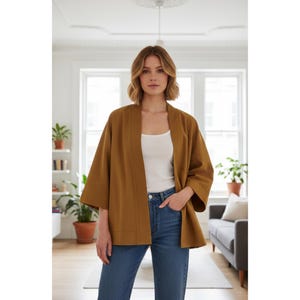 May include: A mustard-colored kimono-style jacket with wide sleeves. The open-front jacket is worn over a white tank top and blue jeans. The model is standing in a well-lit room with a neutral background.