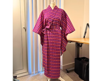 May include: A vibrant, patterned kimono in shades of fuchsia, orange, and purple, displayed on a white mannequin. The kimono features a repeating diamond design and a matching belt. The garment is floor-length and has wide sleeves.