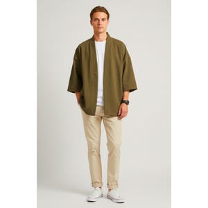 May include: An olive green kimono-style jacket with wide sleeves, worn over a white t-shirt and khaki pants. The outfit is completed with white sneakers and a black watch. The model is standing in front of a white background.