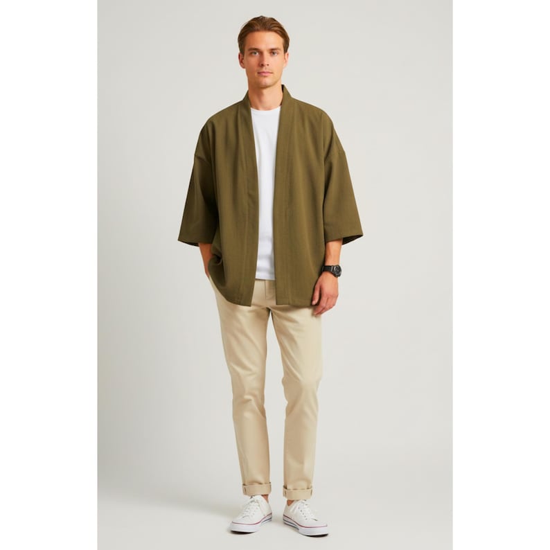 May include: An olive green kimono-style jacket with wide sleeves, worn over a white t-shirt and khaki pants. The outfit is completed with white sneakers and a black watch. The model is standing in front of a white background.