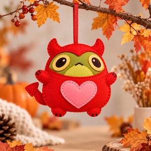 May include: A red felt monster ornament with a green face, large black eyes, and a pink heart patch on its belly. The creature has small horns, a pointed tail, and is suspended by a thin red ribbon, featuring detailed stitching throughout.