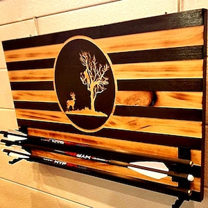May include: Wooden arrow holder with a deer and tree design. The holder features a natural wood grain with black stripes and a circular emblem. Three arrows are held in place by black brackets.