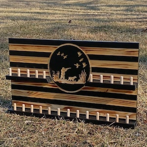 May include: Wooden wall art with a hunting scene. The rectangular piece features alternating black and natural wood stripes. A circular cutout depicts a hunter, dog, and flying ducks. Pegs are placed along the bottom and on two shelves.