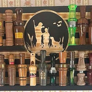 May include: A collection of wooden duck calls and hunting calls displayed on a two-tiered black shelf. The calls vary in size, shape, and wood grain, with some featuring decorative elements. A central wooden plaque depicts a hunting scene.