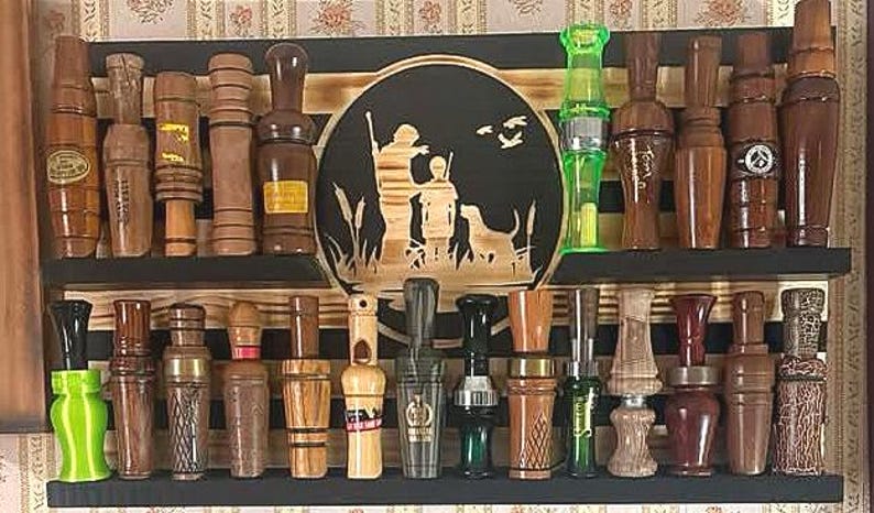 May include: A collection of wooden duck calls and hunting calls displayed on a two-tiered black shelf. The calls vary in size, shape, and wood grain, with some featuring decorative elements. A central wooden plaque depicts a hunting scene.
