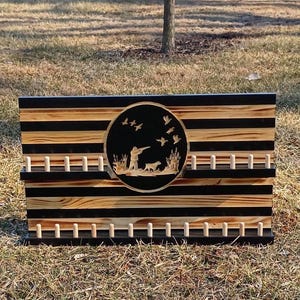May include: Wooden wall art with a hunting scene. The rectangular piece features alternating black and natural wood stripes. A circular emblem in the center depicts a hunter, dog, and flying ducks. Pegs are arranged below and above the emblem.