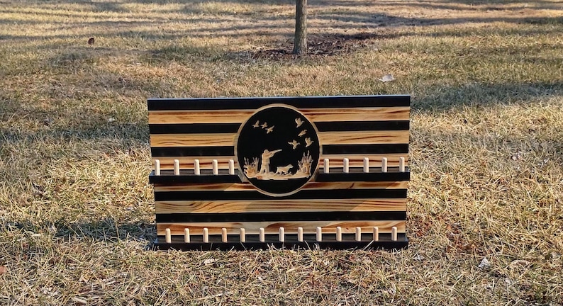 May include: Wooden wall art with a hunting scene. The rectangular piece features alternating black and natural wood stripes. A circular emblem in the center depicts a hunter, dog, and flying ducks. Pegs are arranged below and above the emblem.