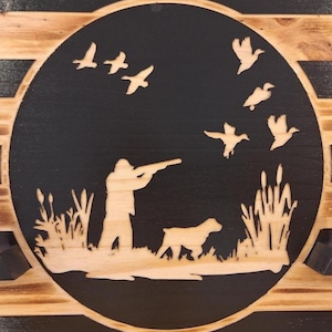 May include: A wooden plaque featuring a hunting scene. A hunter aims a rifle at flying ducks, with a dog at his side. The design is laser-cut into a black circle, set against a light wood background, with reeds and grass.