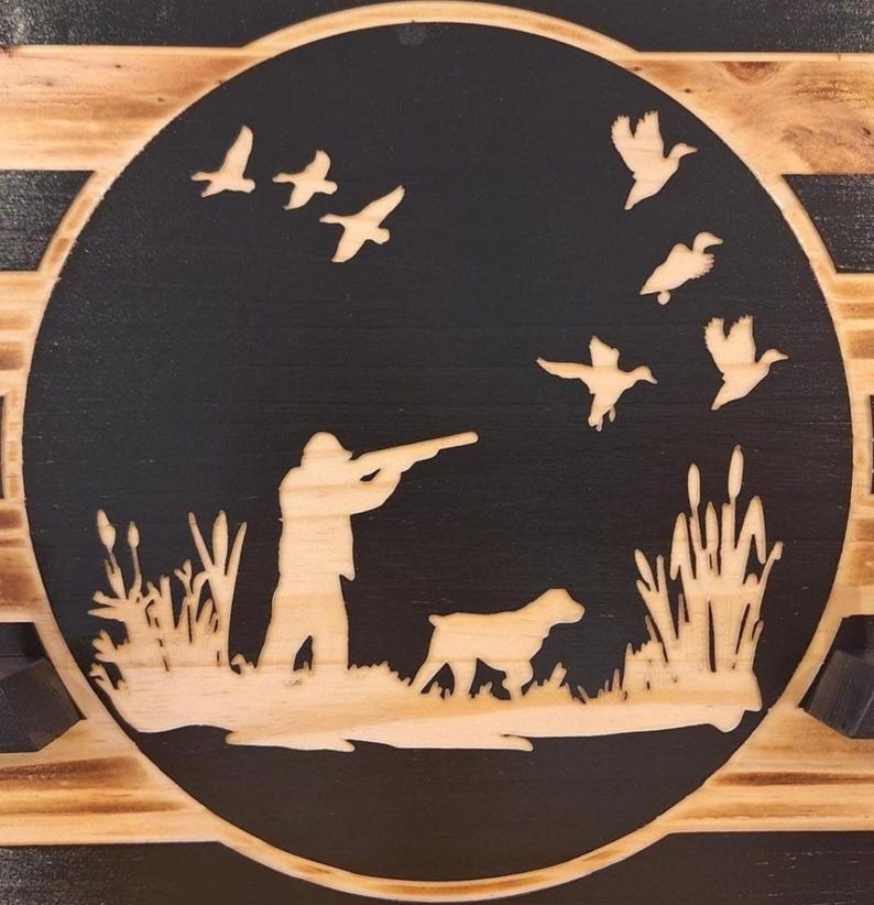May include: A wooden plaque featuring a hunting scene. A hunter aims a rifle at flying ducks, with a dog at his side. The design is laser-cut into a black circle, set against a light wood background, with reeds and grass.