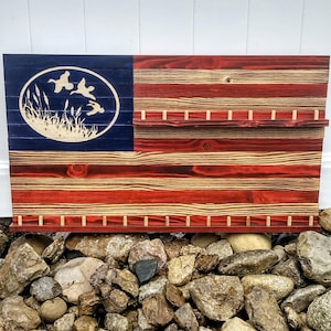 Puede incluir: Una bandera americana de madera con un cantón azul que presenta un óvalo blanco con patos voladores y juncos. La bandera tiene rayas rojas y de madera natural, con clavijas y dos estantes.