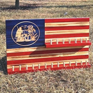 Puede incluir: Bandera americana de madera con rayas rojas y de madera natural. La sección azul presenta un grabado circular de una escena de caza. La bandera tiene dos filas de clavijas para colgar objetos.