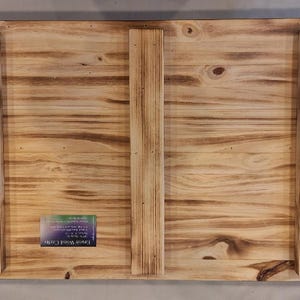 May include: Back of a rectangular wooden sign with a natural wood grain finish. Three vertical wooden support beams are attached with screws. The wood has a burnt, distressed look. A small label is affixed to the lower left corner.