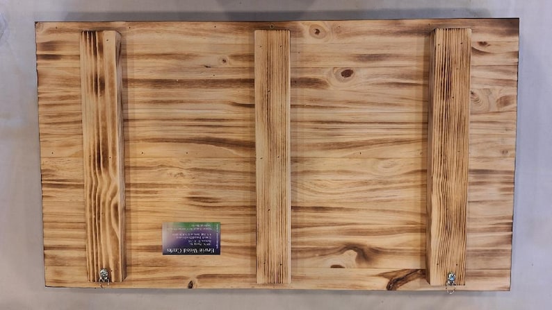 May include: Back of a rectangular wooden sign with a natural wood grain finish. Three vertical wooden support beams are attached with screws. The wood has a burnt, distressed look. A small label is affixed to the lower left corner.