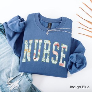 May include: Indigo blue sweatshirt with the word "NURSE" in a floral design. The letters are filled with a pattern of flowers in pink, yellow, and white on a light blue background. A silver necklace is draped across the sweatshirt.