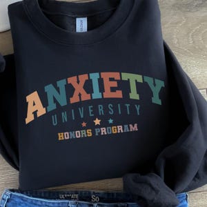 Puede incluir: Una sudadera negra con la frase "ANXIETY UNIVERSITY HONORS PROGRAM" en un diseño retro multicolor. El texto es de color naranja, verde azulado, verde y marrón. Dos estrellas doradas están debajo de la palabra "UNIVERSITY".