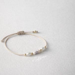 May include: A beige cord bracelet with a sliding knot. It features white, pink, and purple beads, and small gold-coloured beads. The cursive text "@Softmann" is visible. The bracelet is a delicate piece of jewellery.