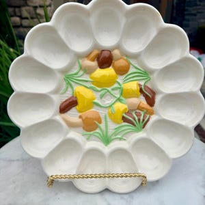 May include: White ceramic deviled egg serving platter with a scalloped edge and a decorative center featuring yellow, brown, and tan mushrooms with green accents. The platter is supported by a gold-toned metal stand.