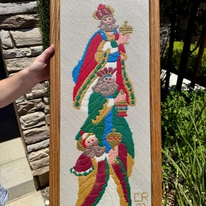 May include: Framed needlepoint art featuring three kings in colorful robes, each holding a crown or chalice. The artwork is set against a neutral background and framed in a light brown wooden frame. The piece is signed "CR 93".