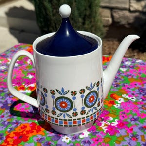May include: A white ceramic teapot featuring a dark blue lid with a spherical knob. The body is decorated with a repeating geometric pattern of orange, light blue, and olive green circles and leaf-like shapes, with a checkered border at the base.