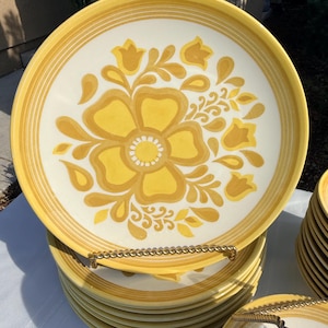 May include: A stack of round ceramic dinner plates featuring a cream-colored center with a large, stylized yellow and gold floral pattern. The plates have a wide yellow rim with concentric circular bands and a smooth, glossy finish throughout.