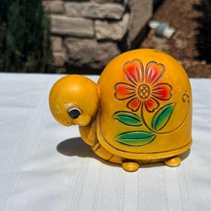 May include: A yellow ceramic turtle coin bank featuring a large embossed flower design on its shell. The flower has orange and red petals with green leaves. The turtle has a smooth yellow finish, a small head, and a coin slot on the top of the shell.