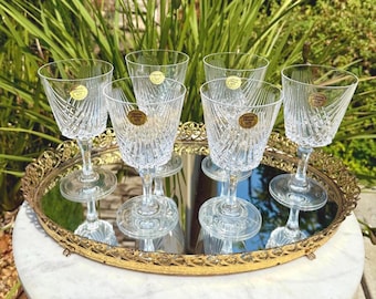 May include: Six clear crystal wine glasses with a textured design, displayed on a gold-rimmed mirrored tray. Each glass has a round base and a decorative label. The glasses are arranged on a white surface with greenery in the background.