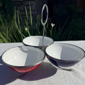 May include: A three-bowl serving dish featuring a central metal handle and a circular frame. The bowls have white interiors and colored exteriors, including one red bowl and one dark blue bowl, all finished with a silver-toned metal rim and base.