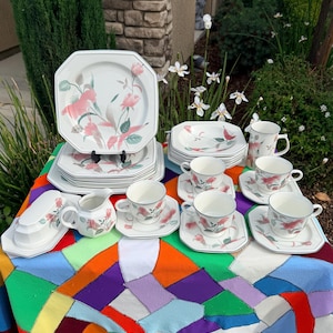 May include: A complete vintage floral dinnerware set. The set includes plates, saucers, cups, a creamer, and a sugar bowl. Each piece features a white background with pink and green floral designs and a silver trim. The set is arranged on a colorful patchwork quilt.