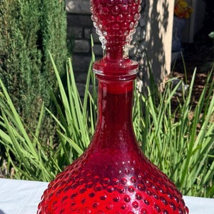 May include: A vibrant ruby red glass decanter featuring a bulbous base and a tall, pointed stopper. Both the body of the decanter and the stopper are covered in a raised hobnail texture, creating a tactile, repeating pattern across the glass surface.