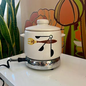 May include: A white ceramic slow cooker with a brown and yellow abstract design. The cooker has a round base and a lid. The base is on a silver metal heating element.